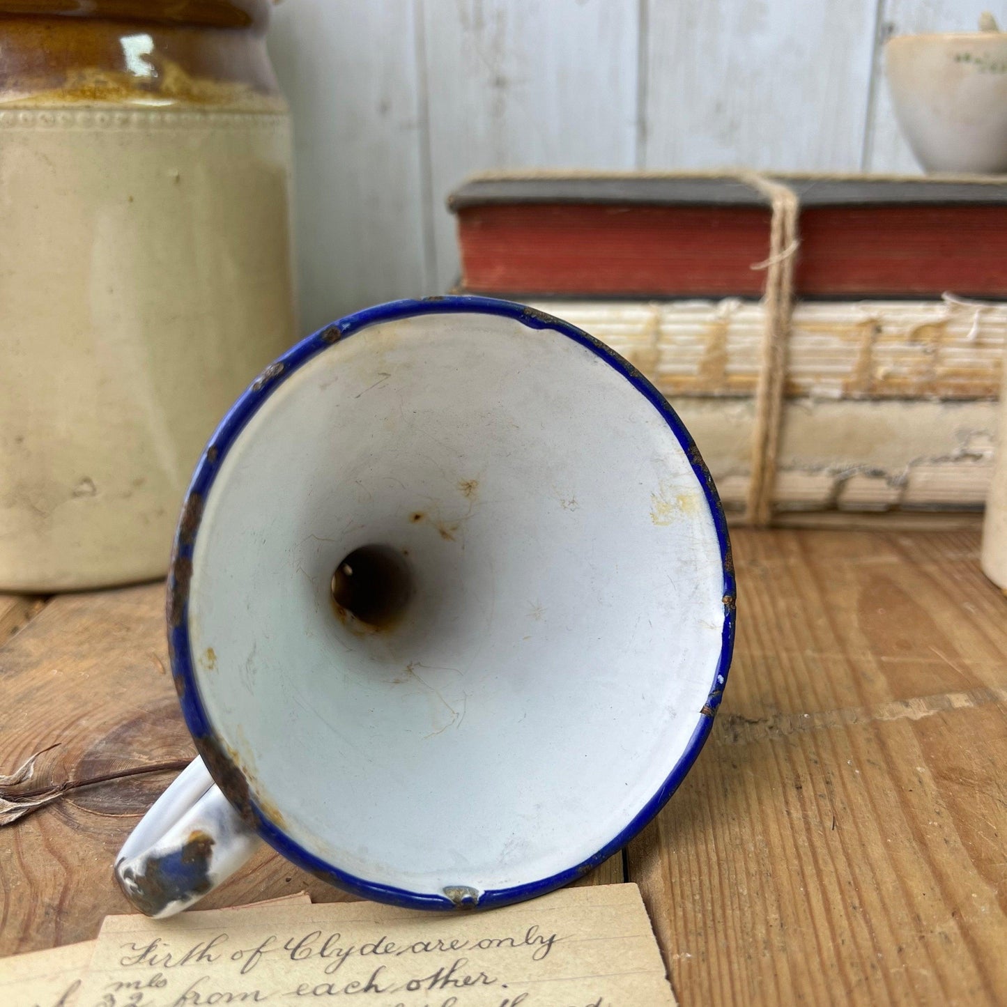 Rusty vintage enamelware funnel with blue rim on wooden surface, old books and pottery in background