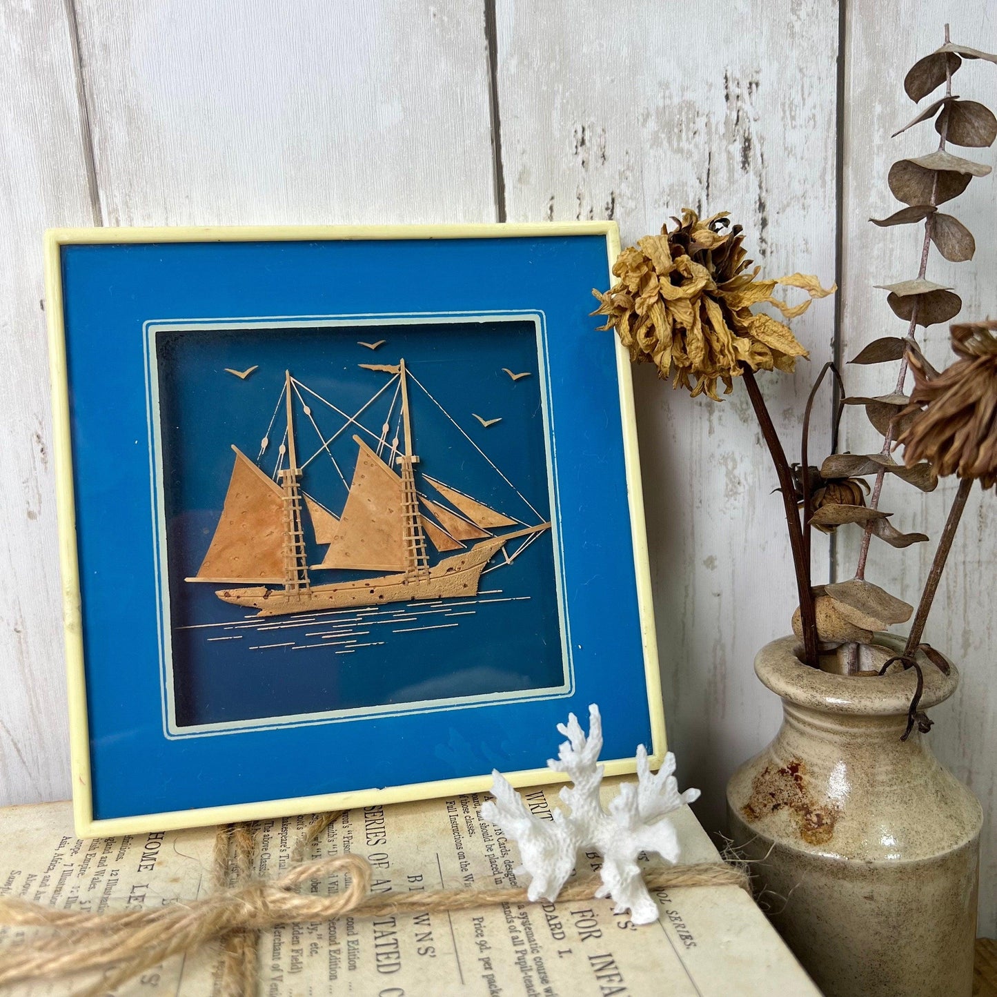 Framed cork sailboat art with blue background, dried flowers in rustic vase, and vintage book