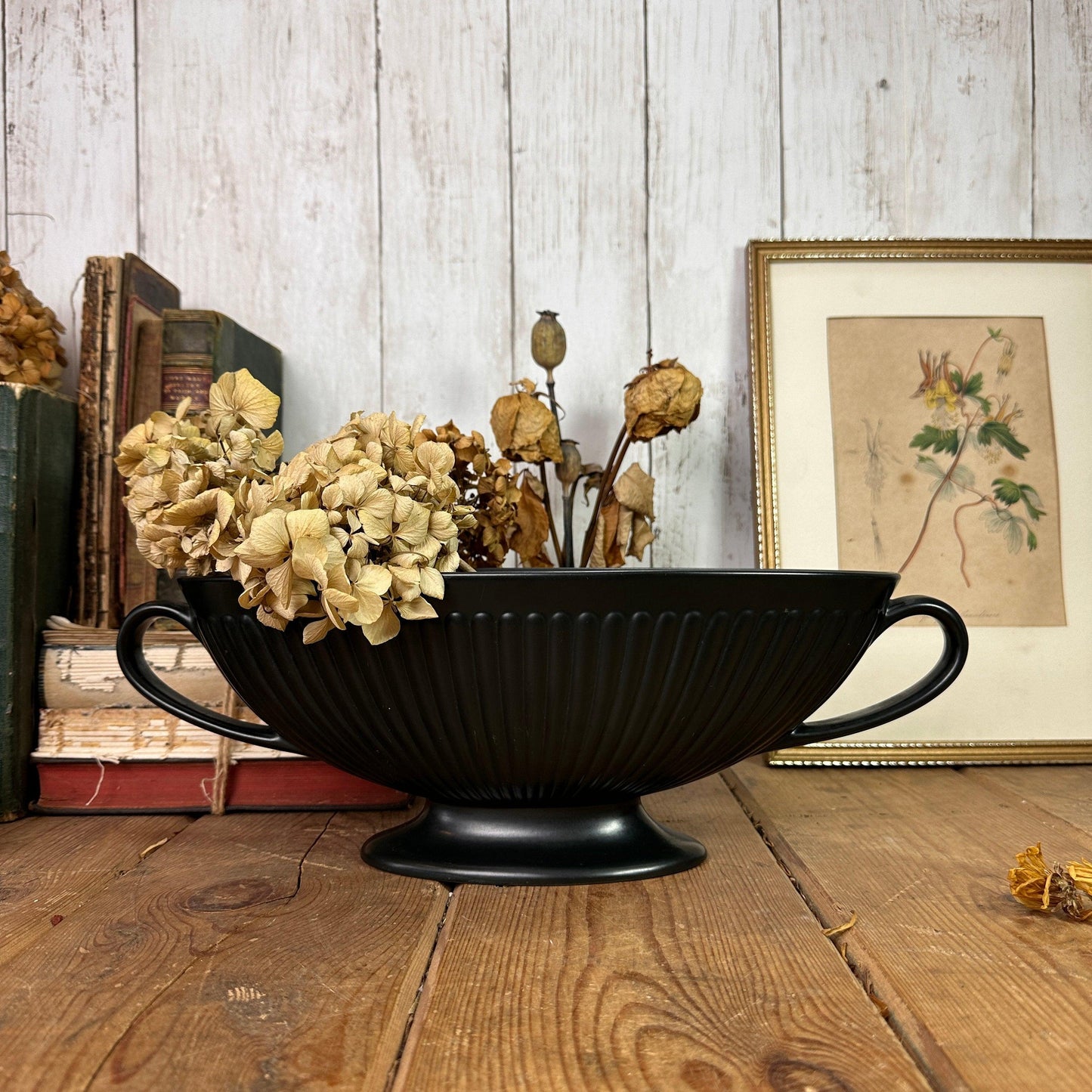 Black matte pedestal bowl holding dried flowers on rustic wooden table with old books and framed botanical print