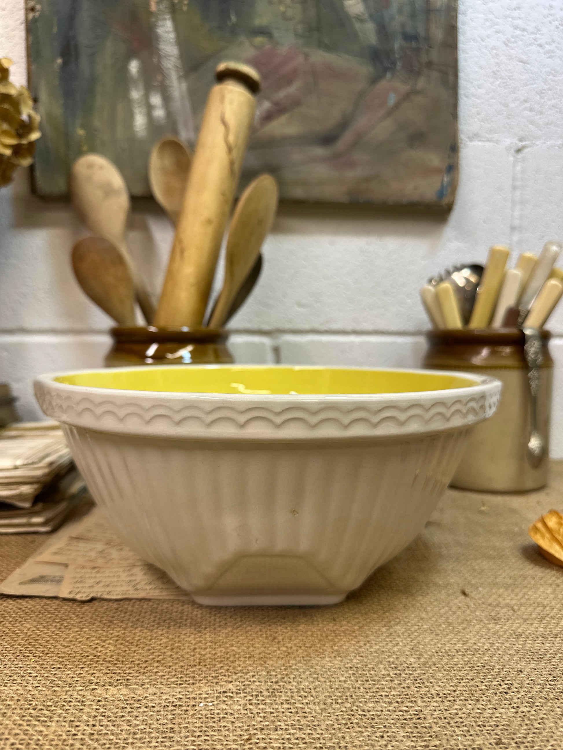 Cream ceramic mixing bowl with yellow interior on burlap mat, wooden spoons and utensils in background